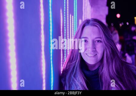 Positive young female in casual clothes looking at camera while standing near glowing neon wall on night street Stock Photo