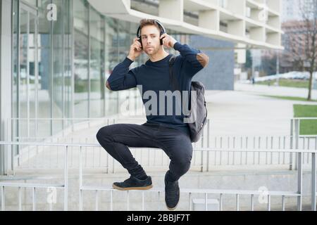 Low angle of modern millennial male in casual outfit with laptop bag listening to music with wireless headphones while sitting on fence on city street Stock Photo