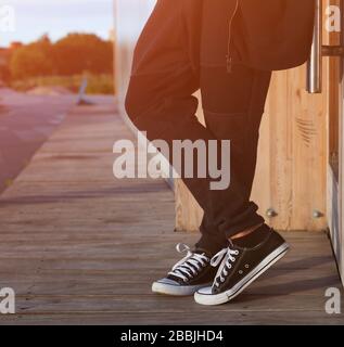 A man in a fashionable black outfit posing at the sunset of a warm summer day in sneakers. Part of the body, close-up. Stock Photo