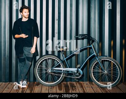 Fun. Night portrait with a flash of a young man in a fashionable black outfit and sneakers posing with a phone and a vintage bicycle. Stock Photo