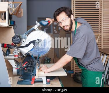 Young hipster man in black overalls by profession carpenter builder saws with a circular saw a wooden board on a wooden table in the workshop Stock Photo