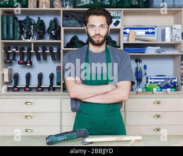 Carpenter master with a beard in work clothes in a carpenter's workshop posing at the desk on the background of equipment Stock Photo