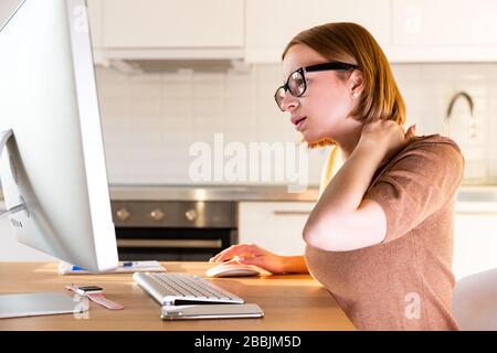 Woman freelancer in beige top suffers from pain in her neck after long work at computer during the period of self-isolation and remote work at home. C