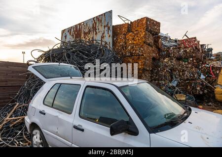 An abandoned car next to a pile of black tubes and cubes of pressed cans in a junkyard Stock Photo