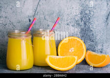 Orange juice in glass jars and fresh oranges on a gray wall background.Close-up. Stock Photo