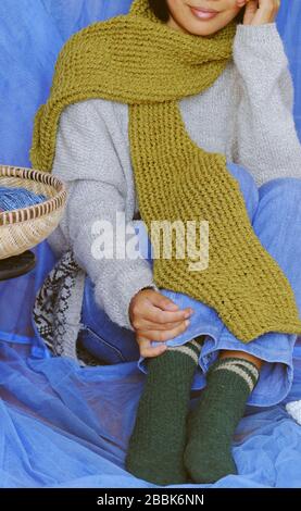 Woman wear blue jeans, white woolen sweater, sock and moss green wool scarf sit with blue background, knitted handmade product to make warm for winter