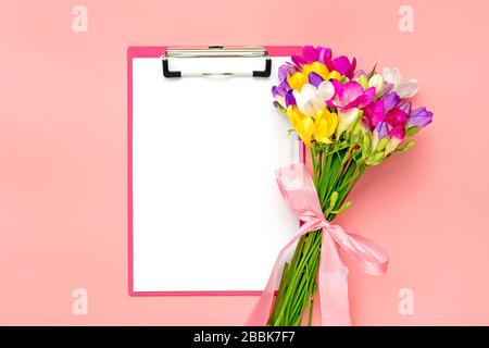 Bouquet of sprig freesia flowers, tablet with white paper isolated on pink background Floral holiday card Top view Flat lay Stock Photo