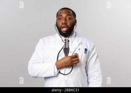 Covid19, hospital check-up and healthcare concept. Funny african-american male doctor in white coat, listening own lungs while breathing, using Stock Photo