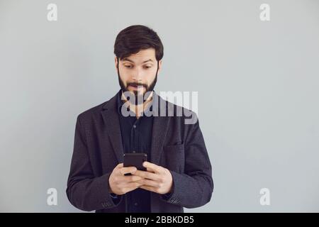 Positive guy in a jacket is typing text in a mobile smiling with on a gray background. Stock Photo