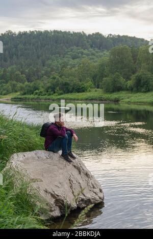 Side view portrait of young man tourist sitting on big stone on narrow forest river bank looking thoughtful and tired. Tourism, hiking, active leisure Stock Photo