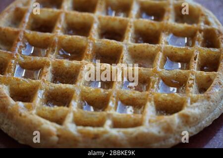 Close-up of one golden waffle with syrup in the indentations reflecting light from a window; grid pattern is slightly diagonal. Full frame. Stock Photo