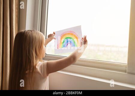 Kid painting rainbow during Covid-19 quarantine at home. Girl near window. Stay at home Social media campaign for coronavirus prevention, let's all be Stock Photo