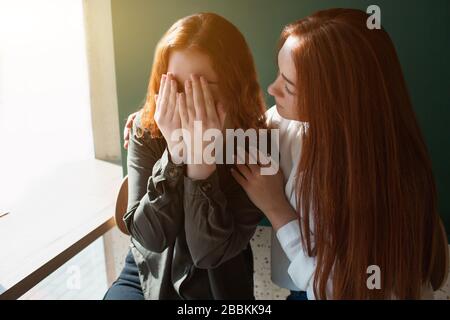 Young woman crying in a cafe covers her face with hands. Female model hugs her friend. Stock Photo