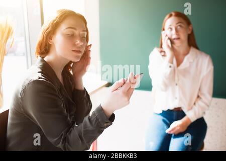 Female model takes a selfie in a cafe and her friend speaks on the phone. Young women are sitting in a cafe. Stock Photo