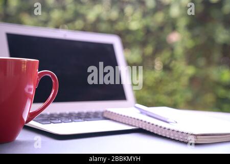 Computer laptop with notepad and coffee in red cup with green nature background. Work remotely or from home. Copy space. Stock Photo