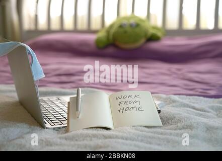 Work from home during coronavirus outbreak. Notepad on computer laptop with surgical face mask on top of bed. Text, work from home written on notepad. Stock Photo