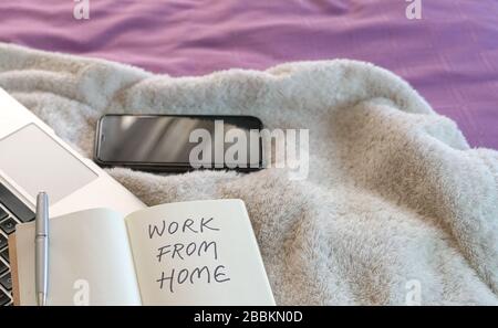 Work from home, text written on notepad. Computer laptop, cellphone and notepad on top of bed in the bedroom. Stock Photo