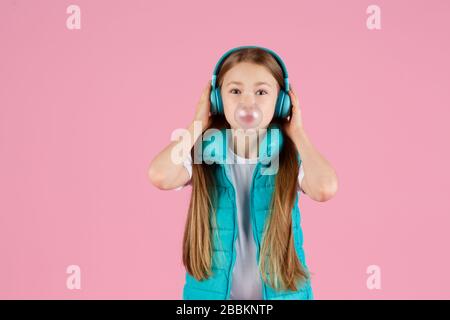 A little girl with headphones explodes pink chewing gum on a pink background. Stock Photo