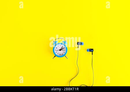Small blue classic-style alarm clock and blue earphones in minimalist style on bright yellow background. Running time. Stock Photo