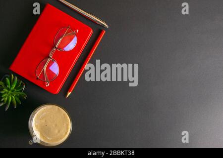 The red notebook stands on a black background, on the notebook there are glasses and next to the notebook, a flower, coffe, pen and a red pencil Stock Photo
