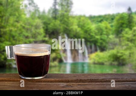 Glass cup of black coffee with waterfall in the forest blur background. View of waterfalls with turquoise water and green trees in Plitvice Lakes Stock Photo