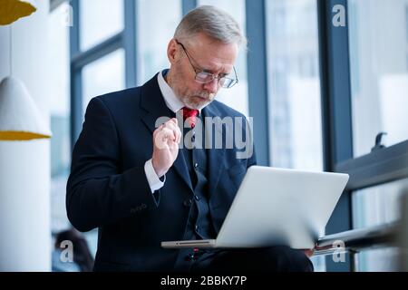 Smiling happy managing director thinks about his successful career development while standing with a laptop in his office near the background of a win Stock Photo
