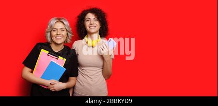 Charming caucasian students with curly hair are posing on a red wall with empty space holding a mobile and some books Stock Photo