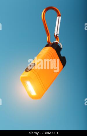 An orange led flashlight with a carabiner glows on a blue background. Stock Photo