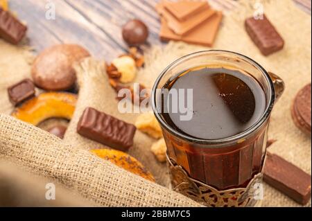 A faceted glass of tea in a vintage Cup holder, pieces of chocolate, bagels and chocolate gingerbread on a background of rough homespun fabric. Close Stock Photo