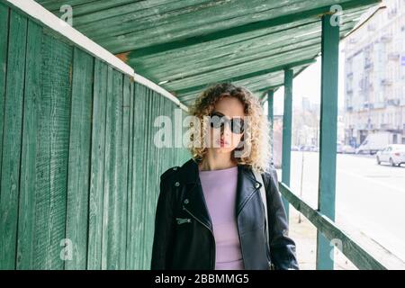 Portrait of beautiful women with curly long hair in black leather jacket and violet dress and black sun glasses in the green building transition Stock Photo