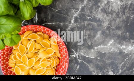 Dry pasta in patterned red bowl on black marble work top with basil Stock Photo