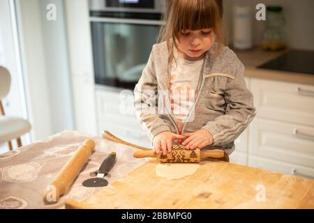 Child girl, 3 years old, in white kitchen, flattening pizza dough with a rolling pin on a wooden board. Lockdown activity idea. Stock Photo