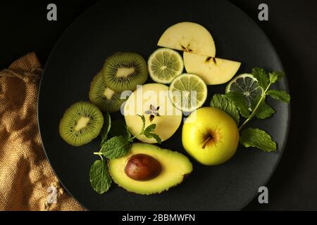 Multiple green fruits, slices of fruits  on round black plate on black background with canvas. Dark dramatic food concept.  Flat lay, top view Stock Photo