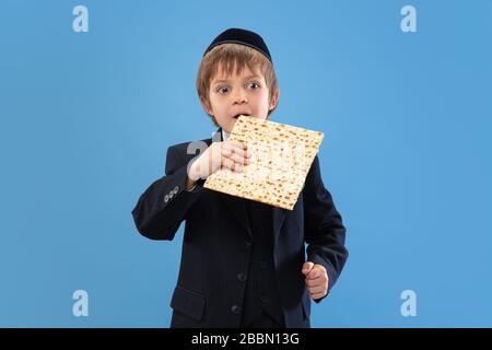 Joyfull eating matzah. Portrait of a young orthodox jewish boy isolated on blue studio background. Purim, business, festival, holiday, childhood, celebration Pesach or Passover, judaism, religion concept. Stock Photo