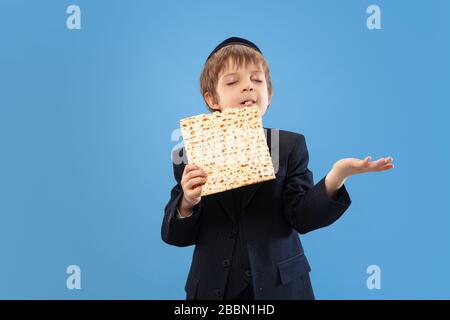 Joyfull eating matzah. Portrait of a young orthodox jewish boy isolated on blue studio background. Purim, business, festival, holiday, childhood, celebration Pesach or Passover, judaism, religion concept. Stock Photo