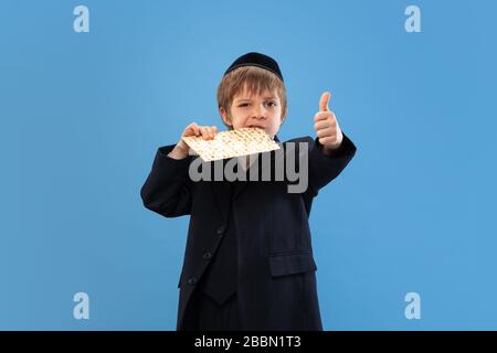 Joyfull eating matzah. Portrait of a young orthodox jewish boy isolated on blue studio background. Purim, business, festival, holiday, childhood, celebration Pesach or Passover, judaism, religion concept. Stock Photo