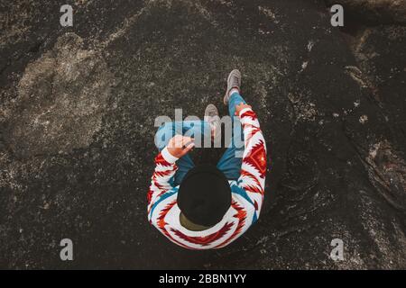 Man relaxing alone travel lifestyle vacations outdoor guy wearing ethnic sweater sitting on dark rocky beach Stock Photo