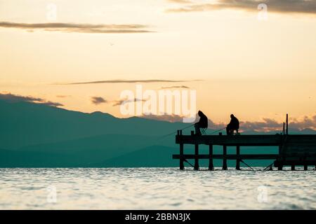 Silhouettes of two fishermen sitting on a dock during sunset. Men fishing while sitting on pier with fishing rods and waiting to catch fish. Stock Photo