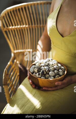 Woman hands holding wooden bowl with quail eggs. Dark background, beautiful natural sun light in kitchen. Easter cooking concept. Stock Photo