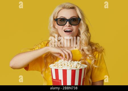 Surprised young blond woman in 3d glasses and yellow shirt eating popcorn, looks shocking movie at the cinema. Isolated on yellow background Stock Photo