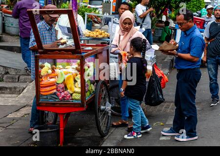 An Indonesian Family Buying Fresh Fruit From A Mobile Street Food Stall, Jakarta, Indonesia. Stock Photo
