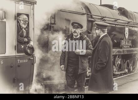 Black & white view railway workers chatting, Kidderminster station platform, Severn Valley heritage railway. Guard leaning out of vintage steam train. Stock Photo