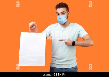 Portrait of joyous buyer, brunette man with surgical medical mask smiling and pointing at packages, shopping bags, surprised about holiday discounts. Stock Photo