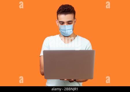 Portrait of cheerful young brunette man with surgical medical mask typing email on laptop and smiling, reading positive message or surfing the web. in Stock Photo