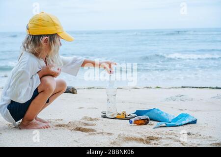 Little boy picks up glass bottle from garbage lying on an ocean shore Stock Photo