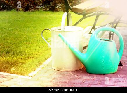 Rubber and tin watering cans near a bench in the summer garden. Sunny morning rays Stock Photo
