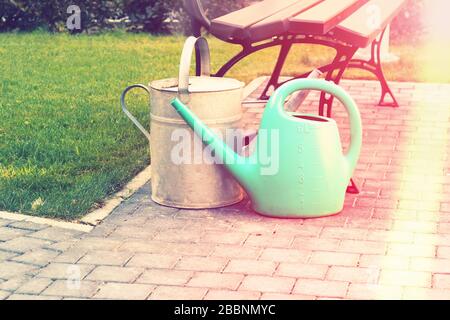 Rubber and tin watering cans near a bench in the summer garden. Sunny morning rays Stock Photo