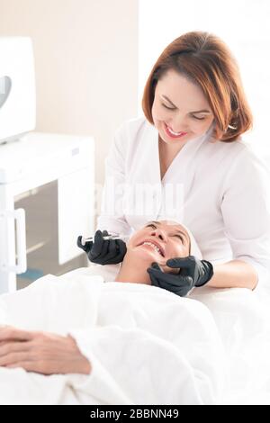 Smiling aesthetic professional in surgical gloves and lab coat injecting filler into face to remove wrinkles of mature woman Stock Photo