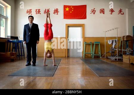Mid-adult man standing beside a teenage girl doing a handstand in a gym. Stock Photo