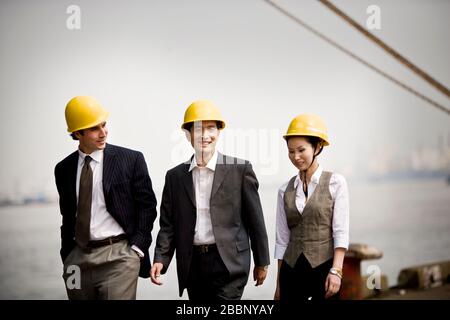 Mid-adult businessman walking with two young adult colleagues along a shipping wharf. Stock Photo